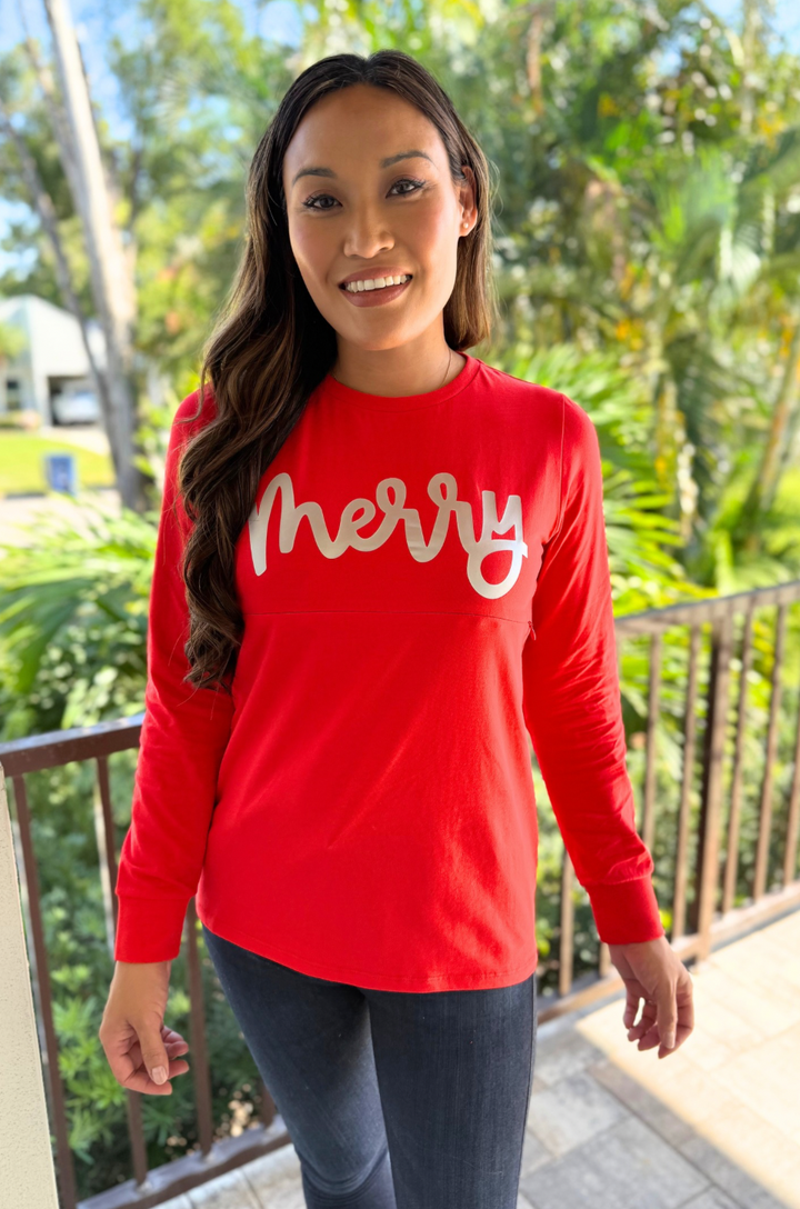 Mom wearing a red 'merry' shirt with nursing access standing outdoors with greenery in the background