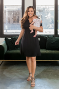 Woman in a black nursing dress holding a baby in a modern living room.