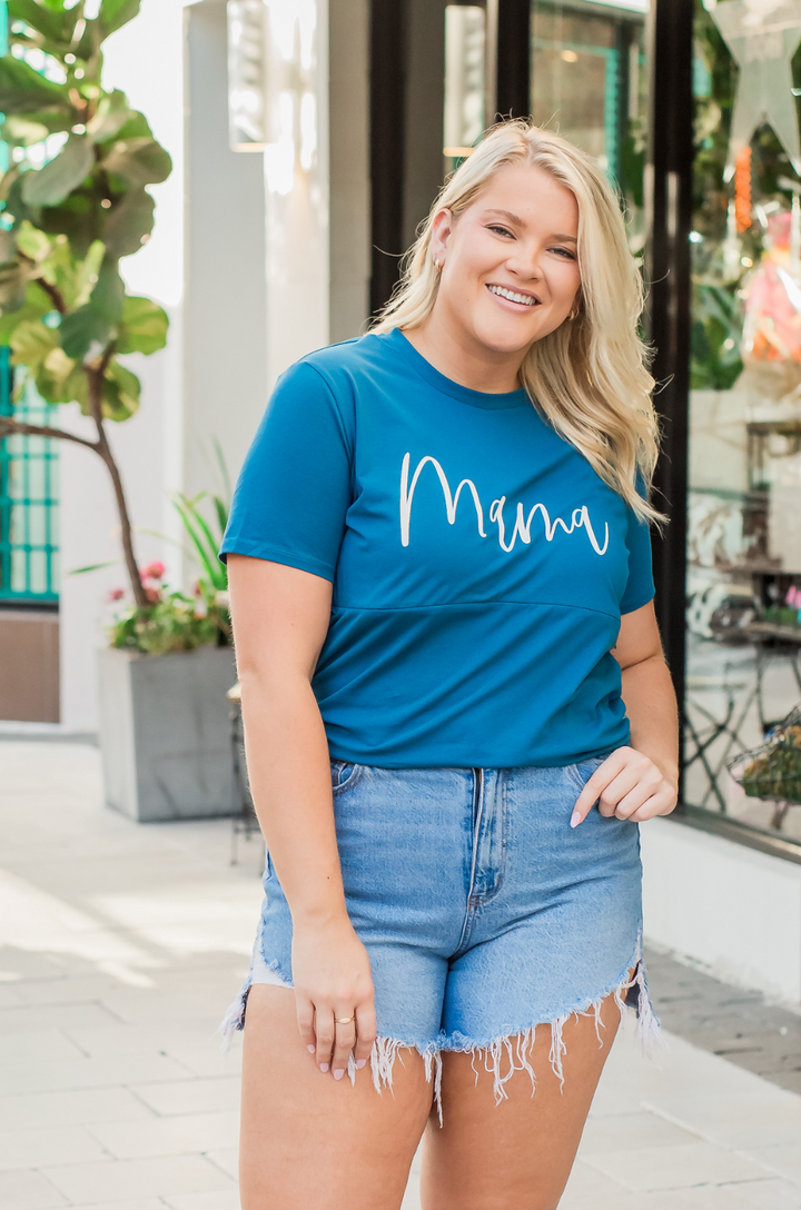 A mom wears the blue Cursive Mama breastfeeding t-shirt paired with shorts for a casual, warm-weather outfit.