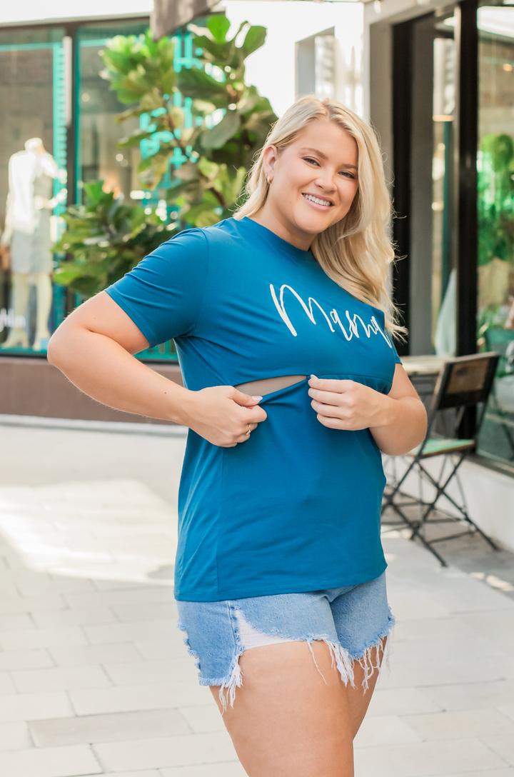 A mom opens the hidden zipper breastfeeding access on the blue Cursive Mama nursing t-shirt while wearing shorts for discreet nursing or pumping.