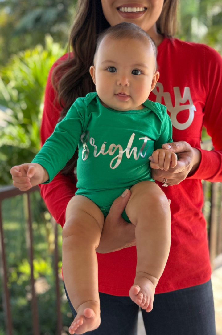 Baby wearing a green onesie with 'Bright' text, held by a person in a red shirt outdoors.