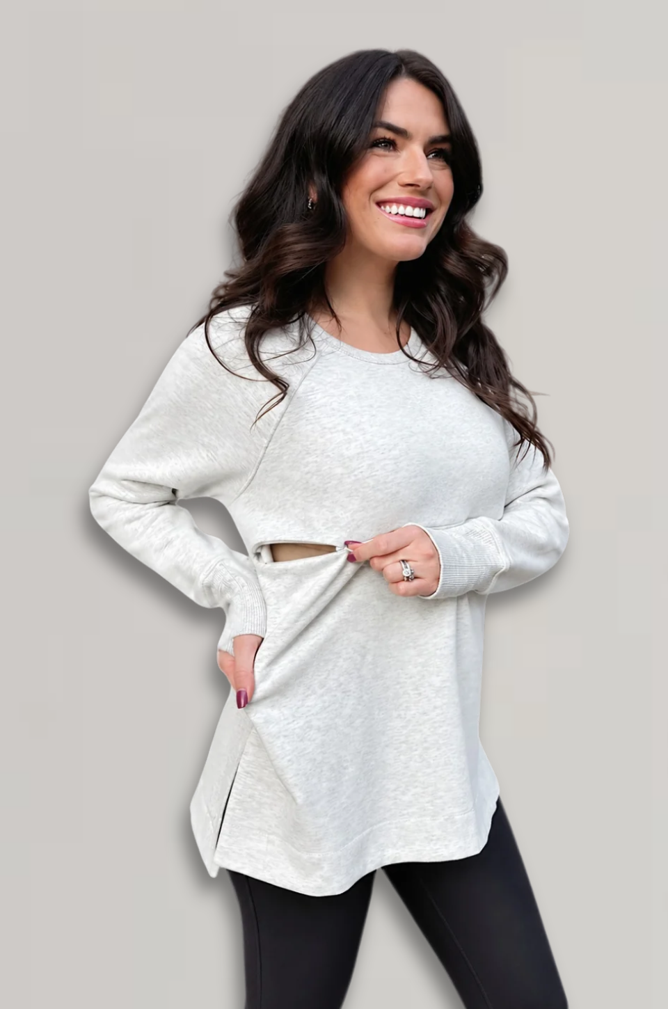 A woman modeling a light heather gray long-sleeve breastfeeding top with a side slit pocket, paired with black leggings, against a solid light gray background.