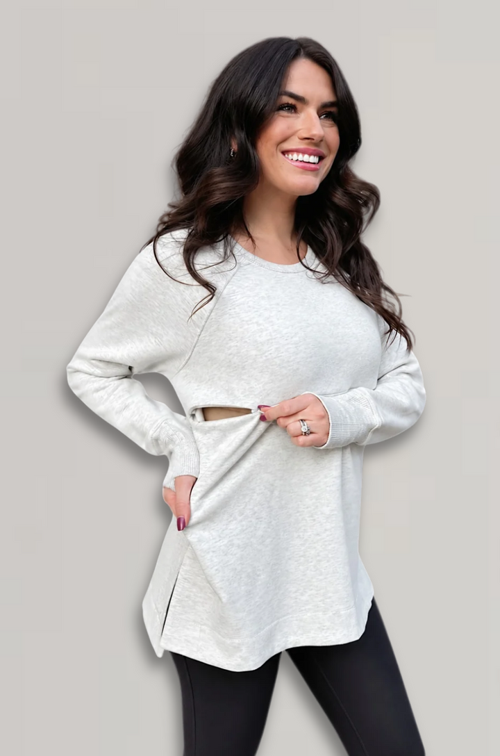 A woman modeling a light heather gray long-sleeve breastfeeding top with a side slit pocket, paired with black leggings, against a solid light gray background.