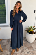 A mom modeling a long-sleeve nursing dress with a V-neckline and gathered waist, standing on a tiled patio next to a white wall and a potted plant.