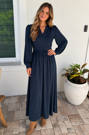 A mom modeling a long-sleeve nursing dress with a V-neckline and gathered waist, standing on a tiled patio next to a white wall and a potted plant.
