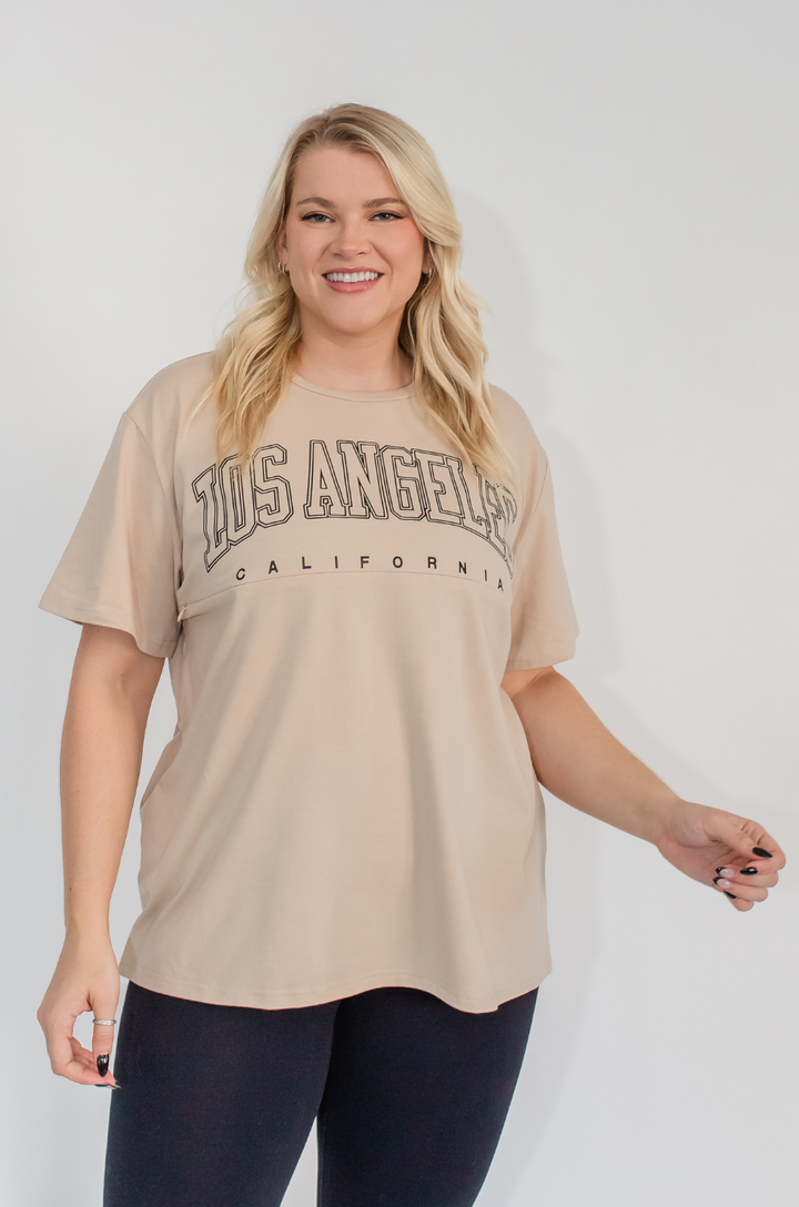 A mom wears the Los Angeles California breastfeeding graphic tan t-shirt with jeans for a casual, everyday outfit.