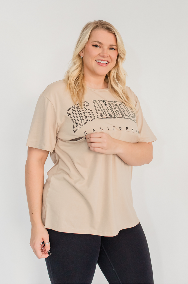 A close-up shows a mom opening the hidden zipper breastfeeding access on the Los Angeles California tan t-shirt for discreet feeding.