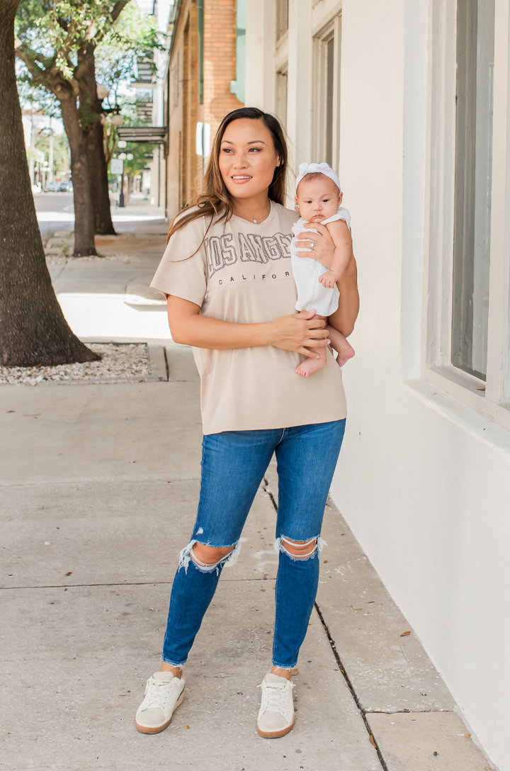 A mom holds her baby while wearing the Los Angeles California breastfeeding graphic tan t-shirt, showing a comfortable real-life breastfeeding moment.