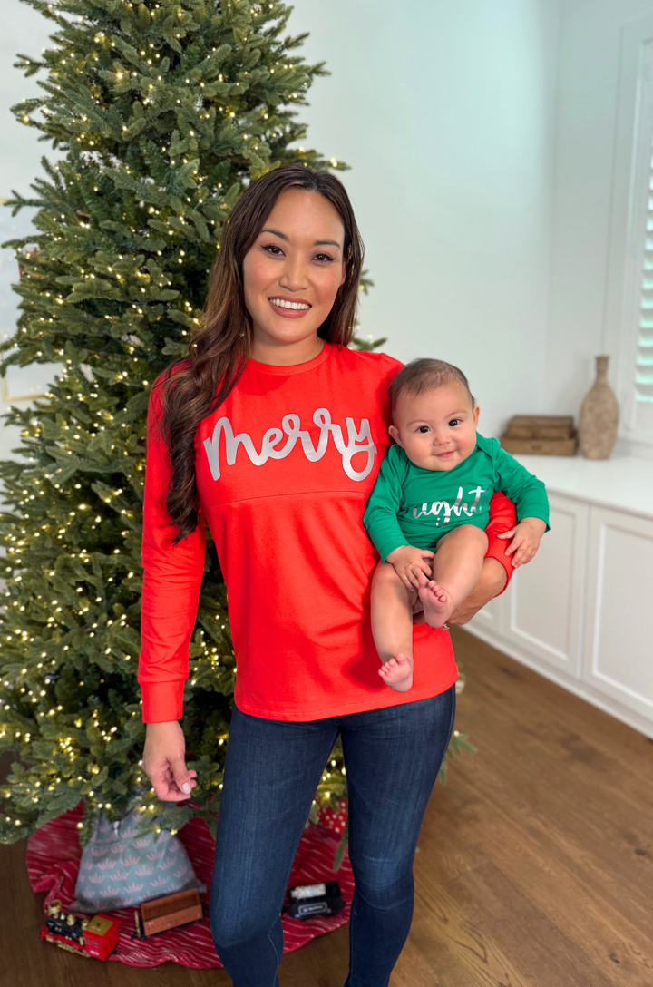 Mom wearing a red shirt that says "Merry" while baby is wearing a green onesie that says "Bright" while standing in front of a Christmas tree. 