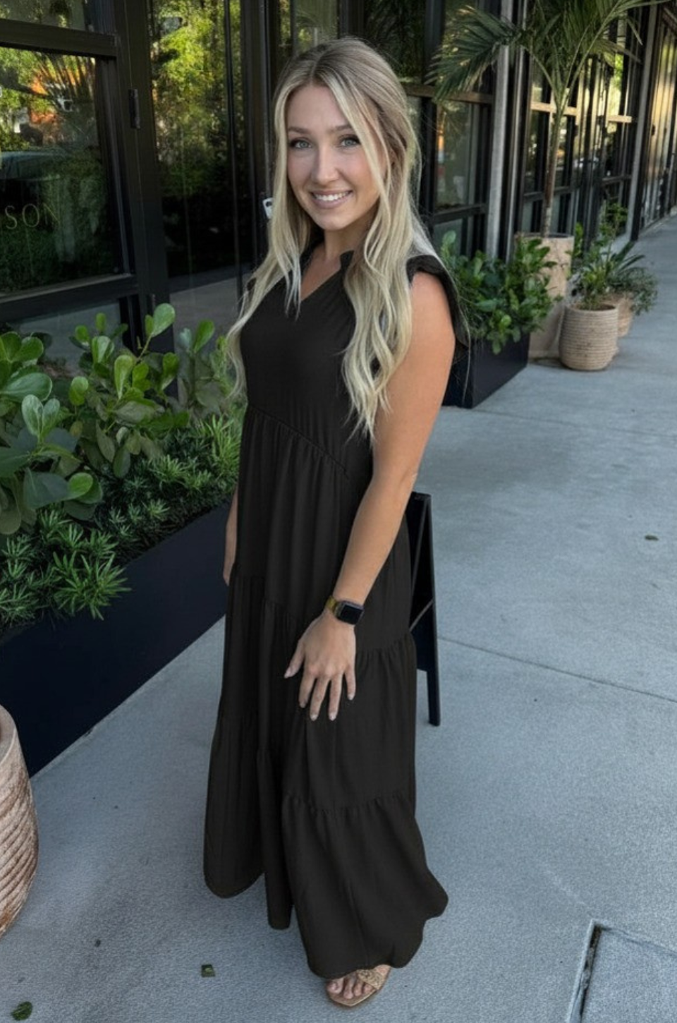 Woman in a black breastfeeding dress standing outdoors with plants and a building in the background
