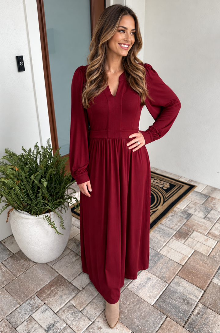 A mom wearing a long-sleeve, deep burgundy maxi dress with nursing access standing on a tiled patio next to a potted fern.