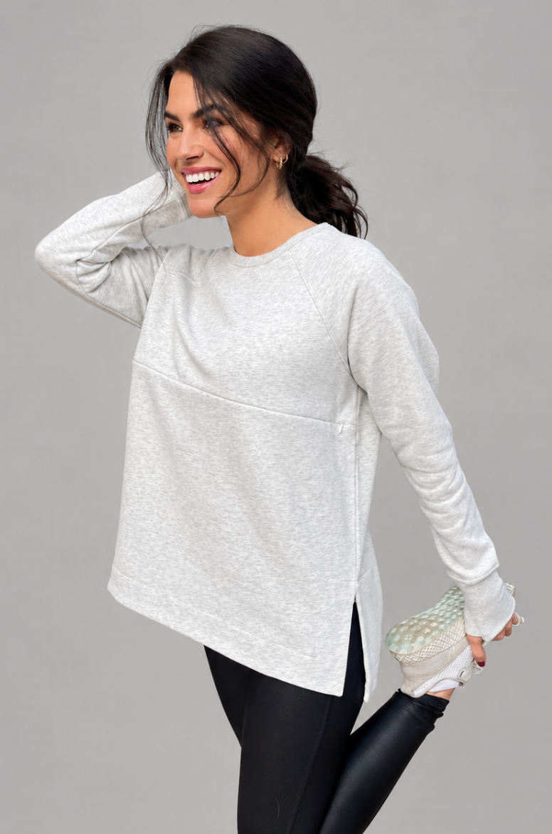 A breastfeeding mom wearing a light gray yoga nursing tunic. posing against a solid gray studio background.