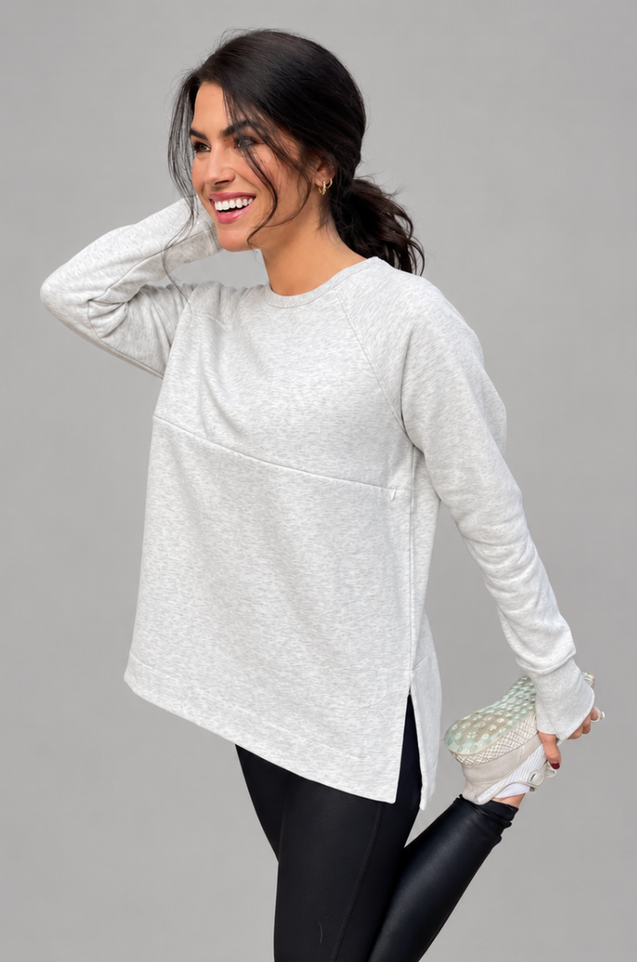A breastfeeding mom wearing a light gray yoga nursing tunic. posing against a solid gray studio background.