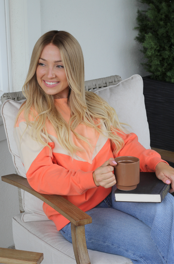 A smiling mom sits in a light beige outdoor armchair, wearing a color-blocked nursing sweater with bright orange, peach, and cream panels. She holds a brown ceramic mug and a black book.
