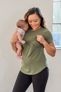 Woman in a breastfeeding green shirt holding a baby against a plain wall.