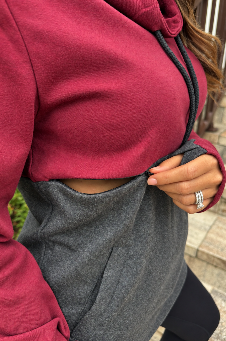A close-up of a person wearing a two-tone hoodie with a bright pink upper section and a heather gray lower section, with a hand showing the zipper access for breastfeeding. 