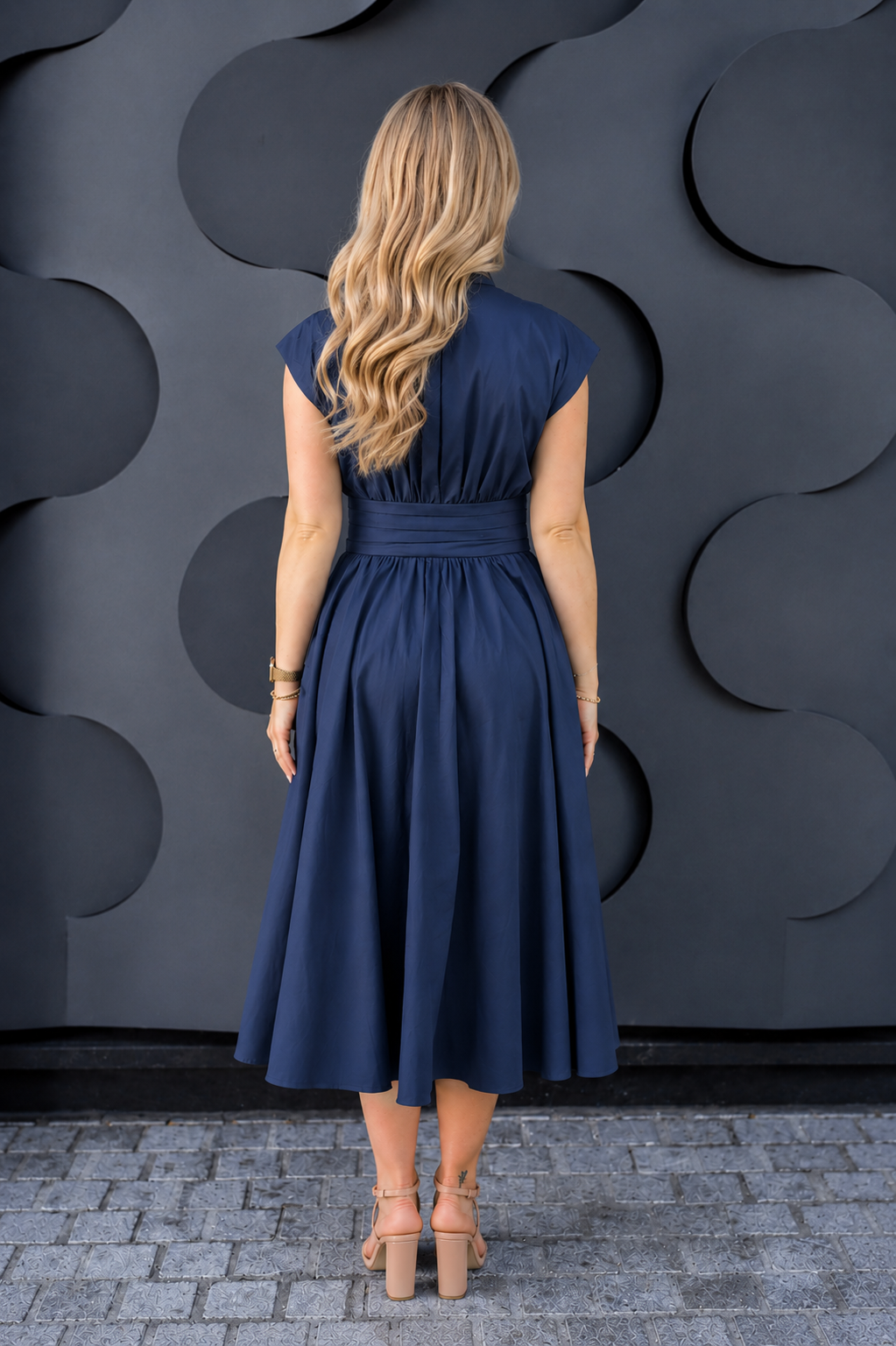 A breastfeeding mom viewed from behind wearing a navy blue midi nursing dress with cap sleeves and a matching waist ti
