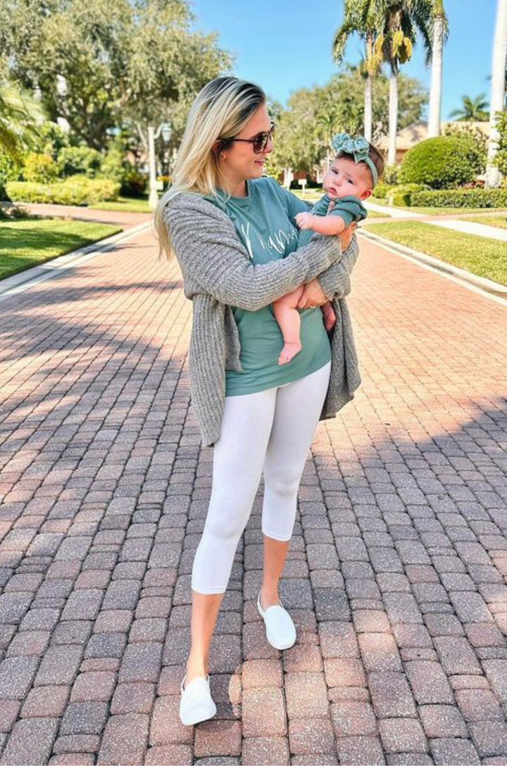 Mother wearing a "mama" shirt, holding her baby dressed in a forest green short sleeve onesie with cursive “baby” text.