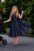 A woman wearing a navy blue midi nursing dress with a tie waist and V-neckline, walking outdoors on a cobblestone path next to a stroller.