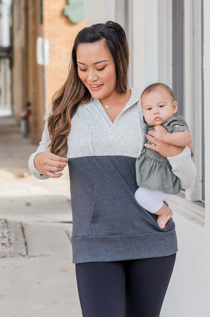 Breastfeeding mom wearing oatmeal waffle nursing hoodie, holding a baby while unzipping the concealed zipper to access nursing opening.