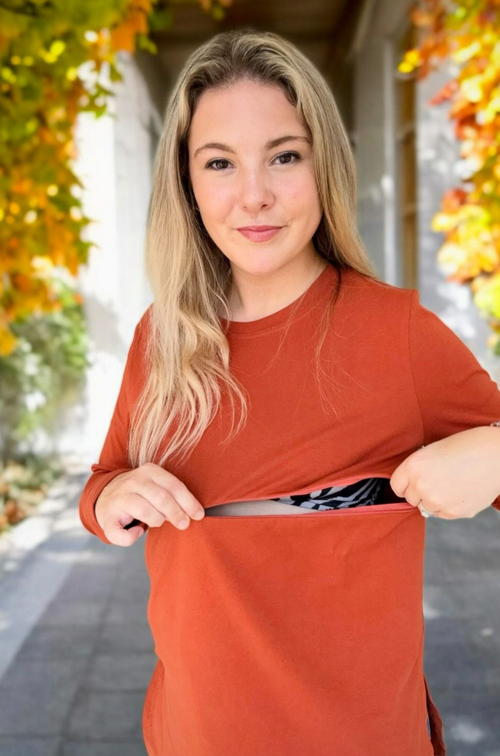Front view of breastfeeding mom wearing rust long sleeve nursing tunic top styled with a white jacket, showing concealed zipper breastfeeding access.