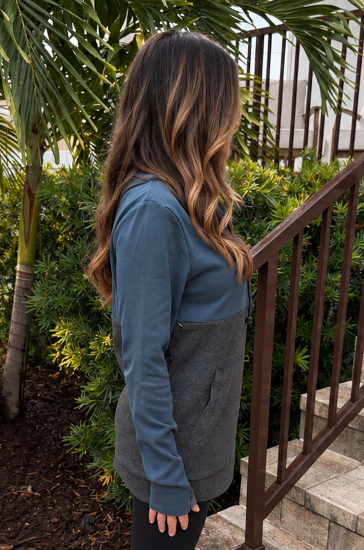 A woman with long wavy brown and blonde-highlight hair is shown from the back, wearing a color-blocked nursing top with a teal upper section and a dark gray lower section, standing outdoors next to a brown metal railing and green plants.